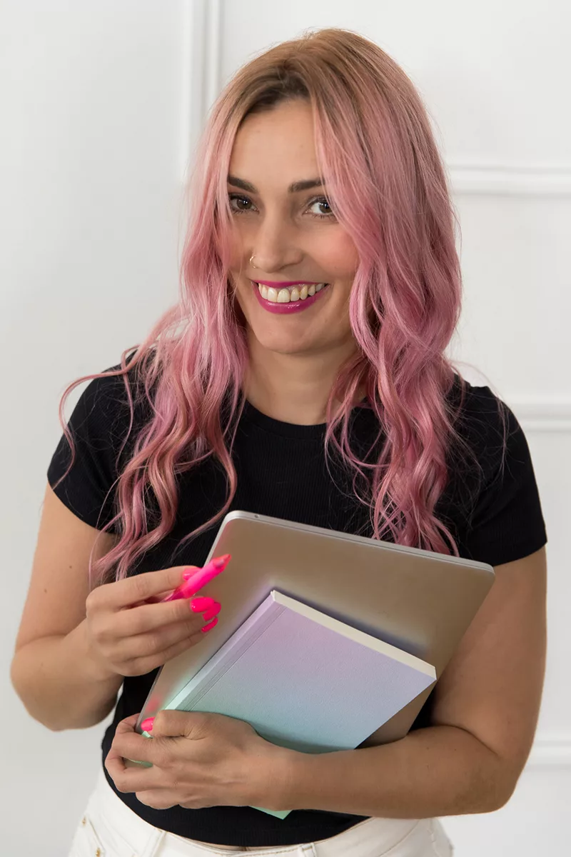 Pip with pink hair and bright neon pink nails, wearing a black t-shirt and white jeans. Smiling and leaning forwards holding a laptop and a notebook. Looking ready to help!